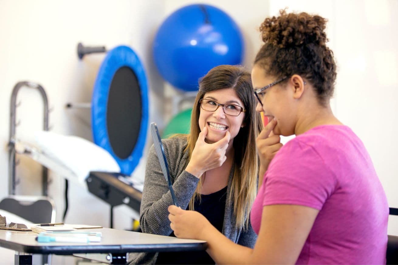 A speech therapist helping an older child work on oral motor skills while holding a mirror